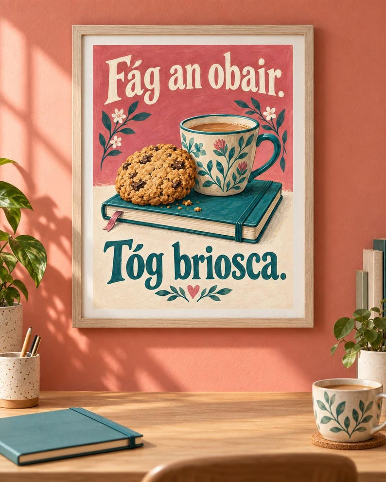 Pu&ograve; includere: Una stampa incorniciata raffigurante un libro verde acqua, un biscotto con gocce di cioccolato e una tazza a motivi floreali su sfondo rosa. Il testo recita F&aacute;g an obair. T&oacute;g briosca. L'illustrazione include piccoli fiori bianchi e un cuore rosa.