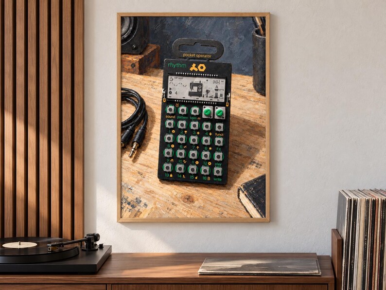May include: A black pocket operator rhythm drum machine featuring a digital LCD screen, a grid of small silver buttons, two green rotary knobs, and yellow text labels. The device has a top handle and displays the text rhythm and pocket operator.