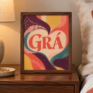 May include: A rectangular framed print featuring the word GR&Aacute; in large red letters centred within a cream-coloured heart shape. The background consists of abstract, flowing shapes in shades of yellow, red, teal, and deep purple with small yellow dashes.