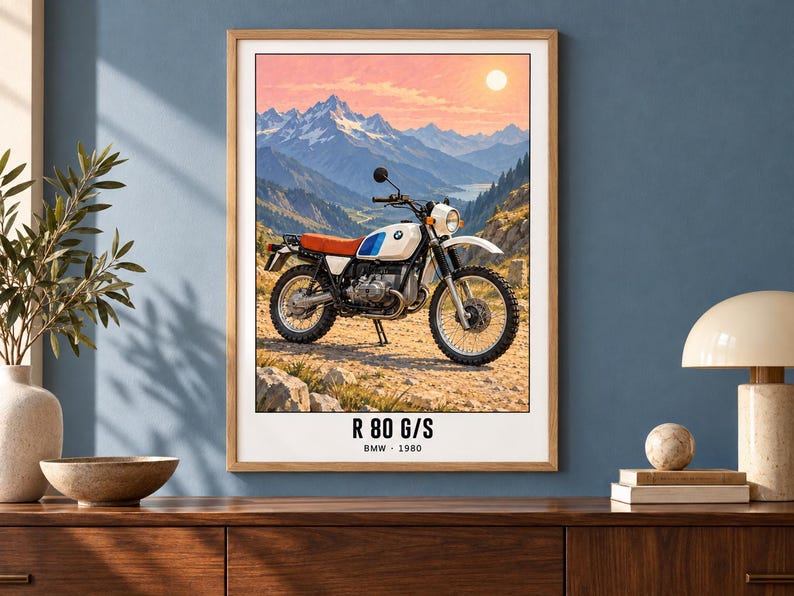 Pu&ograve; includere: Una stampa d'arte incorniciata raffigurante una moto BMW R 80 G/S bianca con sella rossa e dettagli blu su un sentiero sterrato. Lo sfondo mostra una catena montuosa sotto un cielo al tramonto. Il testo in basso riporta R 80 G/S BMW 1980.