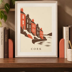 Op de afbeelding: Een ingelijste kunstdruk met een gestileerde illustratie van een rij huizen langs het water in Cork. Het ontwerp gebruikt een palet van cr&egrave;me, donkerbruin en oranje-rood, met het woord CORK in zwarte hoofdletters onderaan in het midden.