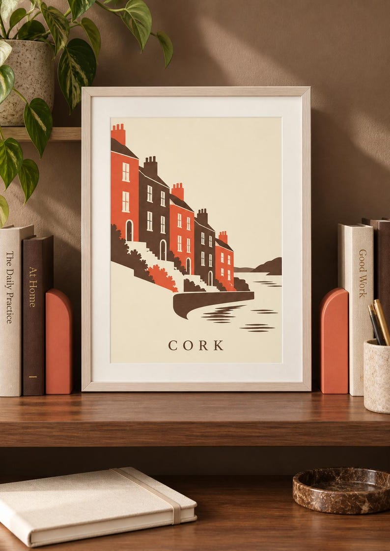 Op de afbeelding: Een ingelijste kunstdruk met een gestileerde illustratie van een rij huizen langs het water in Cork. Het ontwerp gebruikt een palet van cr&egrave;me, donkerbruin en oranje-rood, met het woord CORK in zwarte hoofdletters onderaan in het midden.