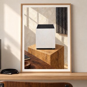 Op de afbeelding: Een ingelijste print met een witte kubusvormige luidspreker met een zwart bovenpaneel, geplaatst op een getextureerd houten blok. Het kunstwerk hangt aan een muur en toont een strakke, moderne esthetiek met neutrale tinten en lijnen.