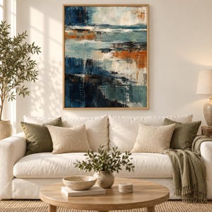 Op de afbeelding: Een ingelijst abstract schilderij met gelaagde horizontale streken in diep marineblauw, taling, wit en gebrand oranje. Het kunstwerk hangt boven een cr&egrave;mekleurige bank met olijfgroene en beige kussens en een gedrapeerde groene plaid.