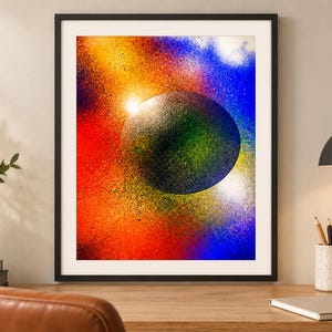 Op de afbeelding: Een ingelijste wandkunstprint met een centrale donkere bol omringd door een levendig, met spuitverf aangebracht nevel-effect in tinten rood, oranje, geel en blauw. De compositie vertoont een getextureerd, gespikkeld uiterlijk met heldere lichtflitsen.