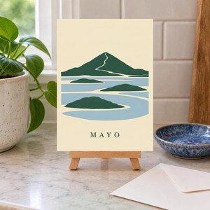 Pu&ograve; includere: Una stampa artistica rettangolare su un piccolo cavalletto in legno con un'illustrazione stilizzata di una montagna e isole in verde scuro e azzurro su sfondo crema. La parola MAYO &egrave; stampata in lettere maiuscole verde scuro in basso.