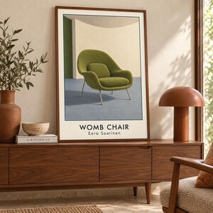 May include: A framed wall art print featuring a green Womb Chair by Eero Saarinen. The illustration shows the chair against a cream and green background with a blue floor. The text Womb Chair Eero Saarinen is printed in black at the bottom.