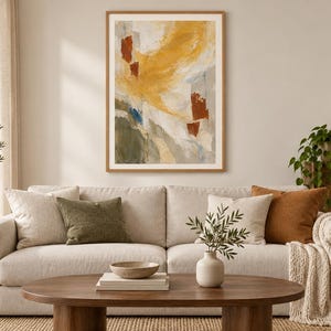 May include: A framed abstract wall art print featuring a composition of mustard yellow, burnt orange, muted green, and grey brushstrokes on a cream background, displayed in a light wood frame hanging above a beige sofa in a living room.