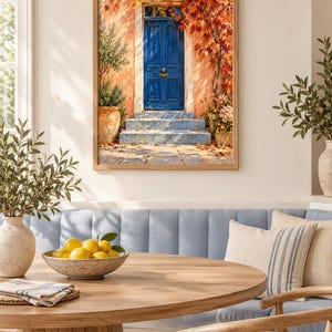 May include: A framed wall art print depicting a vibrant blue door set into a peach-coloured wall, surrounded by autumn foliage in shades of red and orange. The scene includes stone steps, potted plants, and fallen leaves on the ground.