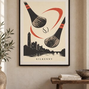 Peut inclure: Une affiche encadr&eacute;e pr&eacute;sentant une illustration stylis&eacute;e de deux crosses de hurling noires et rouges frappant une balle au-dessus d'une silhouette noire du ch&acirc;teau de Kilkenny et d'une rivi&egrave;re. Le mot KILKENNY est inscrit en bas.