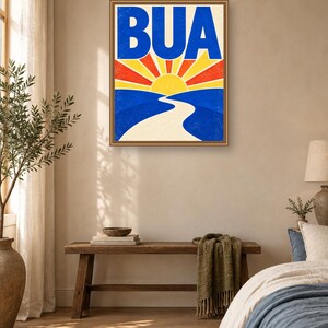 May include: A rectangular wall art print in a thin brown frame featuring the text BUA in large blue letters above a stylized yellow sun rising over a winding white path that cuts through blue hills with red and yellow sunburst rays.