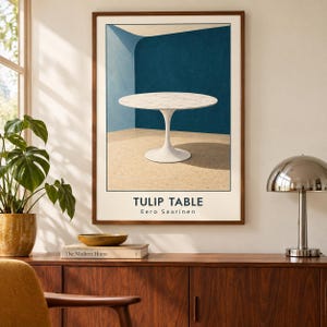 May include: A framed wall art print featuring a white marble Tulip table by Eero Saarinen. The illustration shows the round table against a blue wall and a speckled floor. The text TULIP TABLE and Eero Saarinen is printed at the bottom of the print.