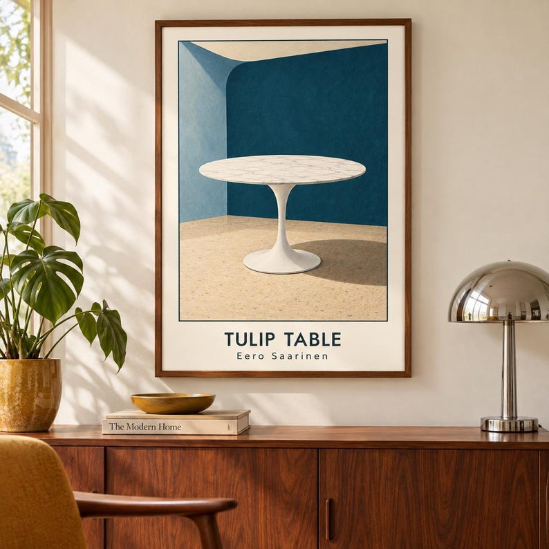 May include: A framed wall art print featuring a white marble Tulip table by Eero Saarinen. The illustration shows the round table against a blue wall and a speckled floor. The text TULIP TABLE and Eero Saarinen is printed at the bottom of the print.