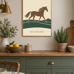 May include: A rectangular wall art print in a thin gold frame featuring a brown horse galloping across a dark green landscape against a cream background. The word KILDARE is printed in dark brown capital letters at the bottom of the print.