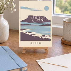 May include: A rectangular art print on a small wooden easel featuring a stylized landscape illustration of a flat-topped mountain, a body of water, and a pale blue sun against a cream background, with the text SLIGO printed at the bottom.