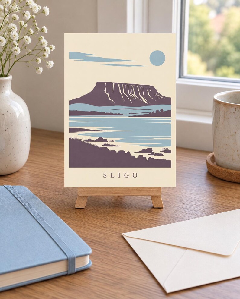 May include: A rectangular art print on a small wooden easel featuring a stylized landscape illustration of a flat-topped mountain, a body of water, and a pale blue sun against a cream background, with the text SLIGO printed at the bottom.