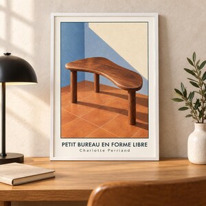 May include: A framed art print featuring a wooden table with a free-form top and three cylindrical legs, set against a blue and cream wall with a tiled floor. The text PETIT BUREAU EN FORME LIBRE Charlotte Perriand is printed at the bottom.