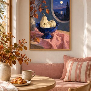 May include: A framed wall art print depicting a still life scene with three white pumpkins in a deep blue pedestal bowl, resting on a pink cloth. The background shows a moonlit coastal landscape viewed through a circular window frame.