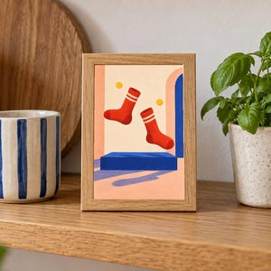 May include: A rectangular wooden frame containing a print of two red socks with white stripes floating above a blue rectangular platform. The background of the print features a light peach wall, a blue archway, and two small yellow circles.