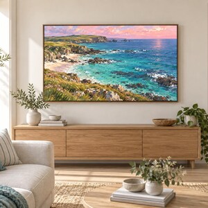 May include: A rectangular landscape painting in a thin wooden frame depicting a coastal scene with turquoise ocean water, rocky cliffs, and green grassy hills under a soft pink and orange sunset sky, mounted on a light beige wall above a wooden console.