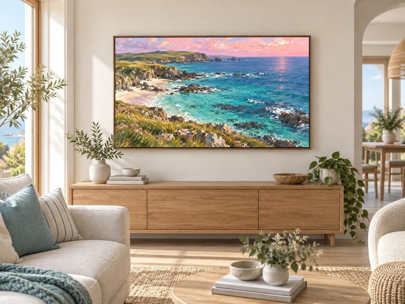 May include: A rectangular landscape painting in a thin wooden frame depicting a coastal scene with turquoise ocean water, rocky cliffs, and green grassy hills under a soft pink and orange sunset sky, mounted on a light beige wall above a wooden console.