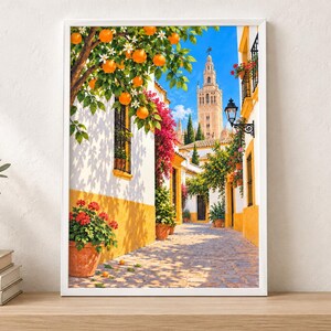 May include: A framed wall art print depicting a sunlit cobblestone street in Seville, Spain. The scene features white buildings with yellow trim, vibrant pink bougainvillea, potted red geraniums, and an orange tree with fruit under a blue sky.