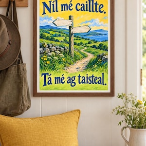 Op de afbeelding: Een ingelijste landschapsprint met een houten wegwijzer op een graspad, glooiende groene heuvels en een blauwe zee. De tekst bovenaan luidt N&iacute;l m&eacute; caillte en de tekst onderaan luidt T&aacute; m&eacute; ag taisteal in donkerblauwe letters.