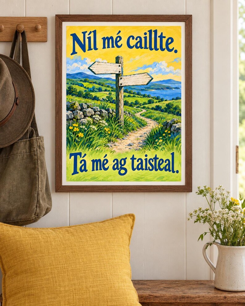 Op de afbeelding: Een ingelijste landschapsprint met een houten wegwijzer op een graspad, glooiende groene heuvels en een blauwe zee. De tekst bovenaan luidt N&iacute;l m&eacute; caillte en de tekst onderaan luidt T&aacute; m&eacute; ag taisteal in donkerblauwe letters.