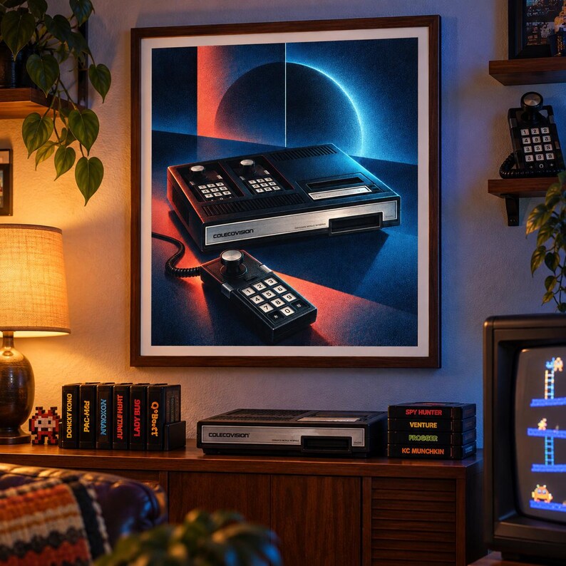 Op de afbeelding: Een ingelijste wandkunstprint met een zwarte ColecoVision-spelconsole en een bedrade controller met numeriek toetsenbord. Het kunstwerk toont een donkere achtergrond met een gloeiend rood en blauw cirkelvormig lichteffect.