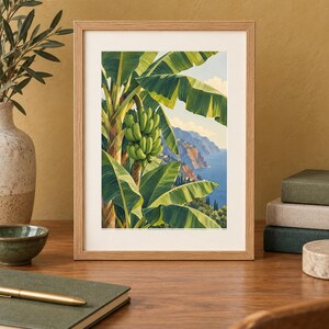 May include: A framed art print featuring a vibrant illustration of a banana tree with a cluster of green bananas, set against a backdrop of a coastal landscape with blue ocean waters, rocky cliffs, and a clear sky with soft white clouds.