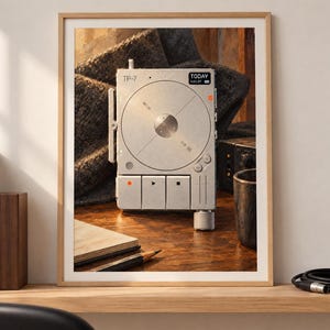 May include: A framed print featuring a silver TP-7 field recorder with a circular central dial, square playback buttons, and a small digital display showing the text TODAY. The device has a metallic finish and is presented in a light wood frame.