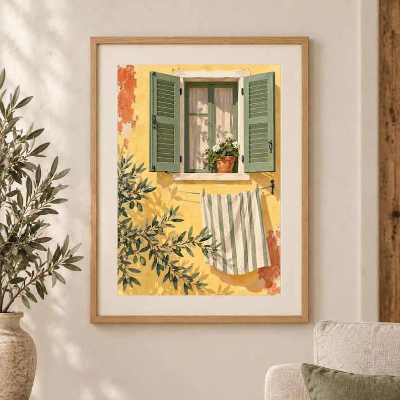 May include: A framed wall art print depicting a yellow stucco wall with an open window featuring green shutters and a potted plant. A white and green striped towel hangs on a clothesline below the window, with olive tree branches in the foreground.