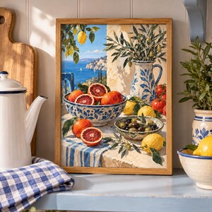 Op de afbeelding: Een ingelijste rechthoekige wandkunstprint met een mediterrane kustsc&egrave;ne met een kom bloedsinaasappels, een kan met olijftakken en een kom olijven op een tafel bedekt met een blauw-wit gestreept kleed, tegen een achtergrond van de zee.