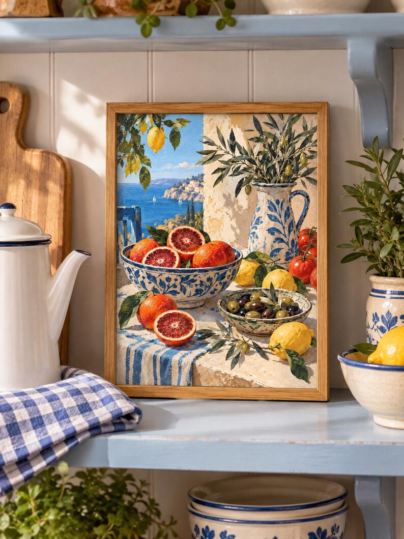 Op de afbeelding: Een ingelijste rechthoekige wandkunstprint met een mediterrane kustsc&egrave;ne met een kom bloedsinaasappels, een kan met olijftakken en een kom olijven op een tafel bedekt met een blauw-wit gestreept kleed, tegen een achtergrond van de zee.