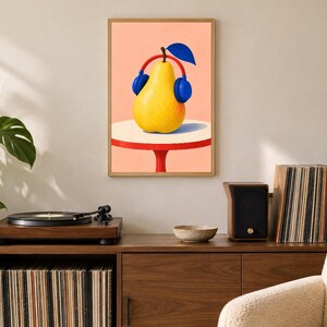 May include: A framed wall art print featuring a yellow pear wearing blue headphones with a red headband. The pear sits on a white circular table with a red base against a solid peach background. The illustration uses bold colours and clean lines.
