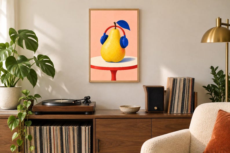 May include: A framed wall art print featuring a yellow pear wearing blue headphones with a red headband. The pear sits on a white circular table with a red base against a solid peach background. The illustration uses bold colours and clean lines.