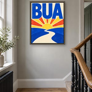 May include: A rectangular framed wall art print featuring the word BUA in large blue letters at the top. Below the text is a stylized graphic of a yellow sun rising over blue hills with a winding cream-colored path leading toward the horizon.