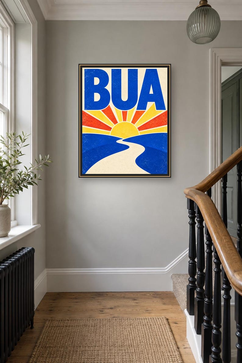May include: A rectangular framed wall art print featuring the word BUA in large blue letters at the top. Below the text is a stylized graphic of a yellow sun rising over blue hills with a winding cream-colored path leading toward the horizon.