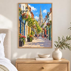 Op de afbeelding: Een ingelijste wandkunstprint met een levendig Europees straatbeeld met kleurrijke winkelpuien, hangende bloemenmanden en een hoge stenen kerktoren in de verte onder een helderblauwe lucht. Mensen lopen over het geplaveide pad.
