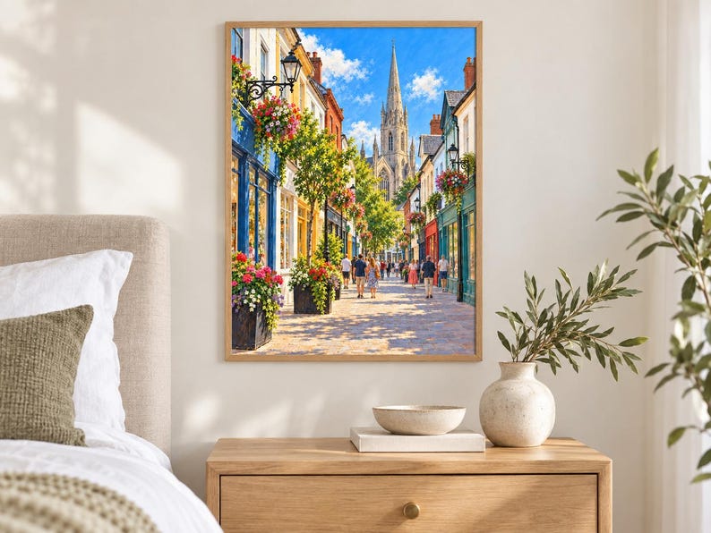 Op de afbeelding: Een ingelijste wandkunstprint met een levendig Europees straatbeeld met kleurrijke winkelpuien, hangende bloemenmanden en een hoge stenen kerktoren in de verte onder een helderblauwe lucht. Mensen lopen over het geplaveide pad.
