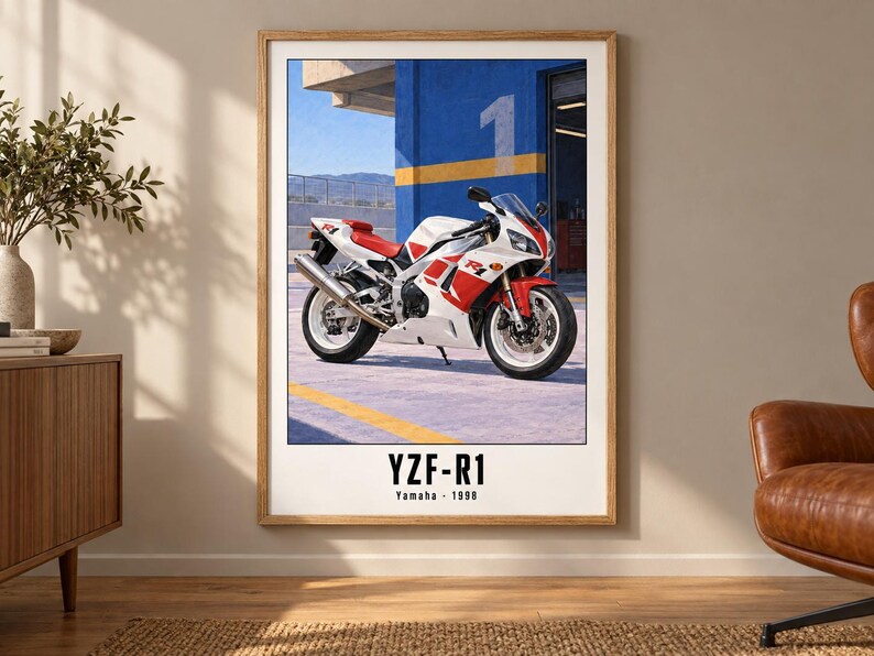 May include: A framed wall art print featuring a white and red Yamaha YZF-R1 motorcycle parked in front of a blue building with a yellow stripe and the number one. The text YZF-R1 and Yamaha 1998 is printed in black at the bottom of the white poster.