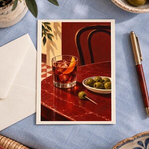 May include: A rectangular art print featuring a dark red cocktail in a glass with ice and an orange peel garnish, placed on a deep red table next to a small white bowl of green olives and a single olive on a toothpick, with a chair in the background.