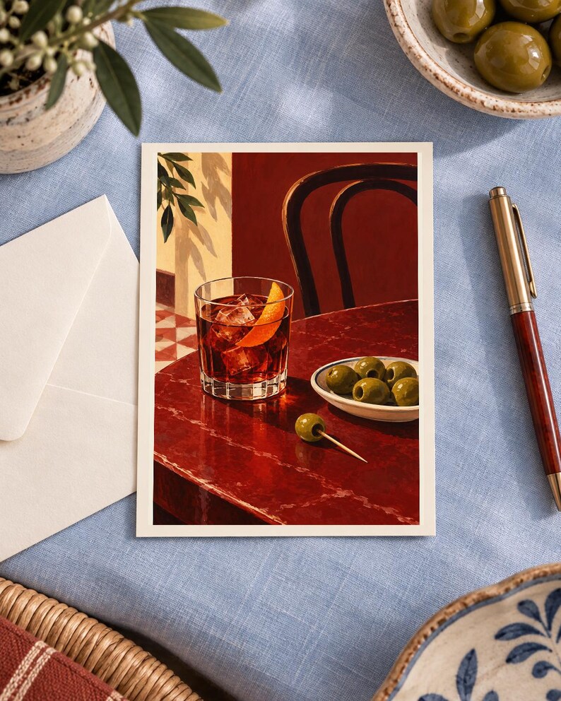 May include: A rectangular art print featuring a dark red cocktail in a glass with ice and an orange peel garnish, placed on a deep red table next to a small white bowl of green olives and a single olive on a toothpick, with a chair in the background.