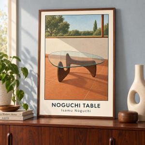 May include: A framed art print featuring a Noguchi table with a glass top and dark wood base, set on a terracotta patio. The text NOGUCHI TABLE Isamu Noguchi is printed at the bottom. The print is displayed in a dark wood frame on a wooden surface.