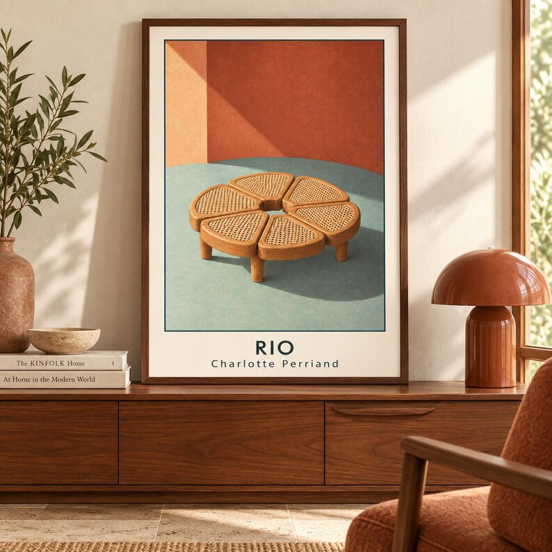 May include: A framed art print featuring a circular wooden table with a woven cane top composed of six wedge-shaped segments. The print displays the text RIO and Charlotte Perriand at the bottom, set against a background of terracotta and teal.