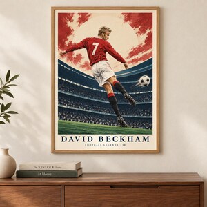May include: A framed poster featuring a soccer player wearing a red jersey with the number 7 and white shorts, kicking a soccer ball in a stadium. The text DAVID BECKHAM FOOTBALL LEGENDS 10 is printed at the bottom against a red and white sky background.
