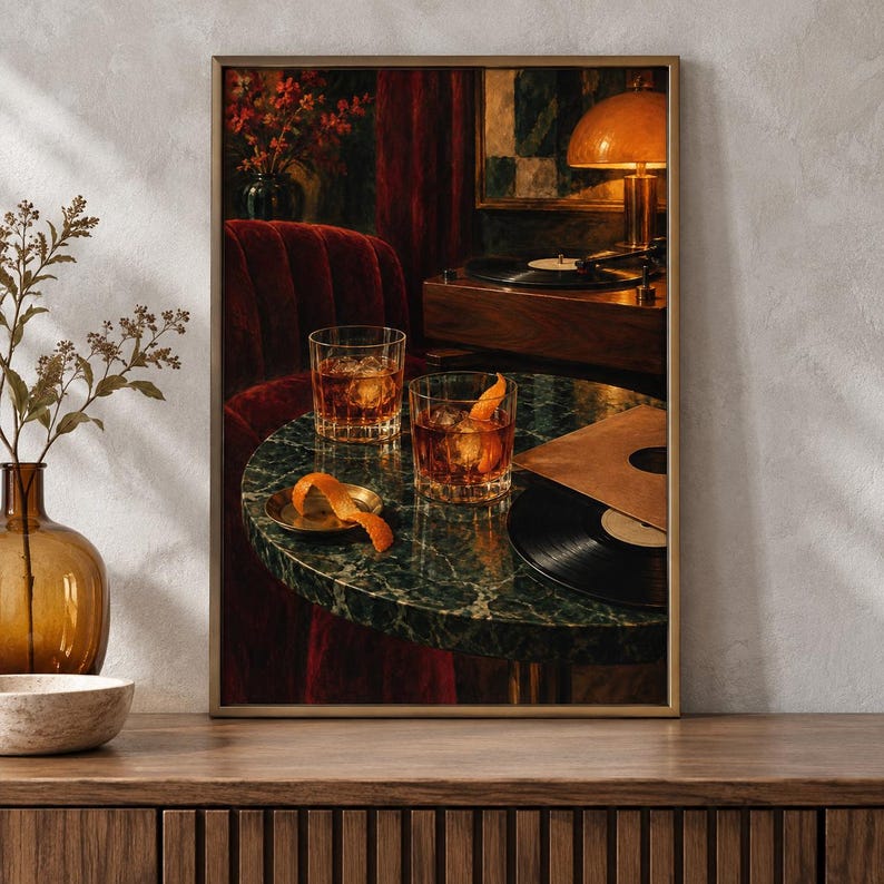 May include: A framed wall art print depicting a dark, moody interior scene with two glasses of amber-colored liquid garnished with orange peels on a green marble table. A vinyl record, a record player, and a warm lamp are visible in the background.