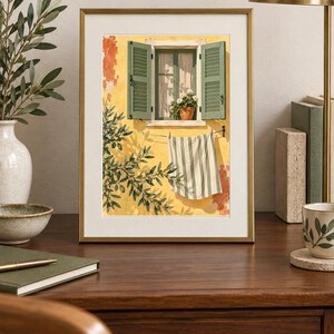 May include: A framed art print featuring a yellow wall with an open window, green shutters, and a potted plant. A striped towel hangs on a clothesline below the window, with olive tree branches casting shadows across the textured yellow surface.
