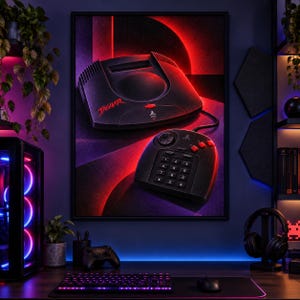 K&ouml;nnte beinhalten: Ein gerahmter Wandkunstdruck, der eine schwarze Atari Jaguar Videospielkonsole und den dazugeh&ouml;rigen Controller zeigt. Die Konsole tr&auml;gt das rote Jaguar-Logo und das Atari-Emblem. Das Bild ist vor einem dunklen Hintergrund mit rotem Licht.