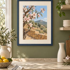May include: A rectangular wall art print in a dark blue frame featuring a landscape painting of a blooming almond tree with light pink flowers, a stone wall, and a distant mountain under a clear blue sky, displayed on a solid olive green wall.